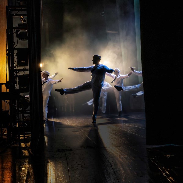 Flera dansare i vita sjömanskostymer rör sig på en scen. Bilden är tagen från sidan av scenen. 