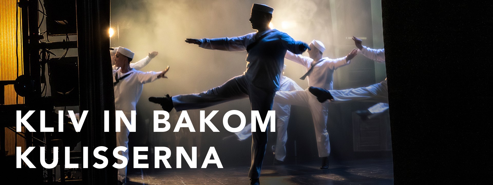 Flera dansare i vita sjömanskostymer rör sig på en scen. Bilden är tagen från sidan av scenen. 