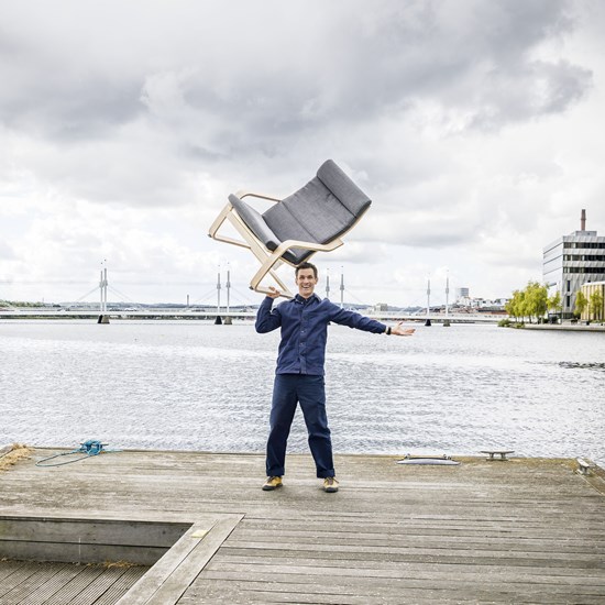 En blåklädd, glad man står på en brygga vid en sjö och balanserar en grå fåtölj i luften. 