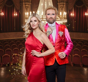 Affischbild för julkonserten En Fridfull Jul med Emmi Christensson och Rickard Söderberg. En man och en kvinna i finkläder står i en upplyst salong med kristallkrona, röda stolar och draperier.