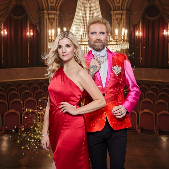 Affischbild för julkonserten En Fridfull Jul med Emmi Christensson och Rickard Söderberg. En man och en kvinna i finkläder står i en upplyst salong med kristallkrona, röda stolar och draperier.
